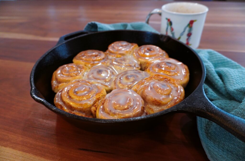 iced cinnamon rolls in a cast iron skillet next to a mug and teal towel