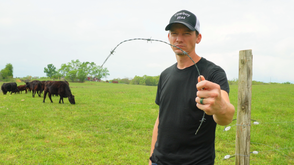 Man in black shirt with ball cap holding a piece of barbed wire fence