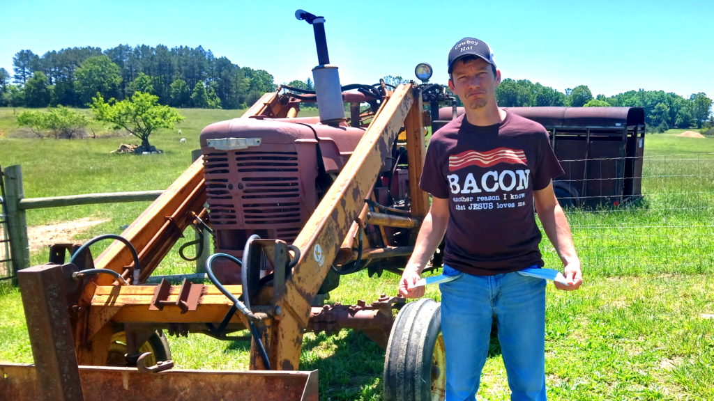 Make in brown shirt and blue jeans holding his pockets standing by an old tractor