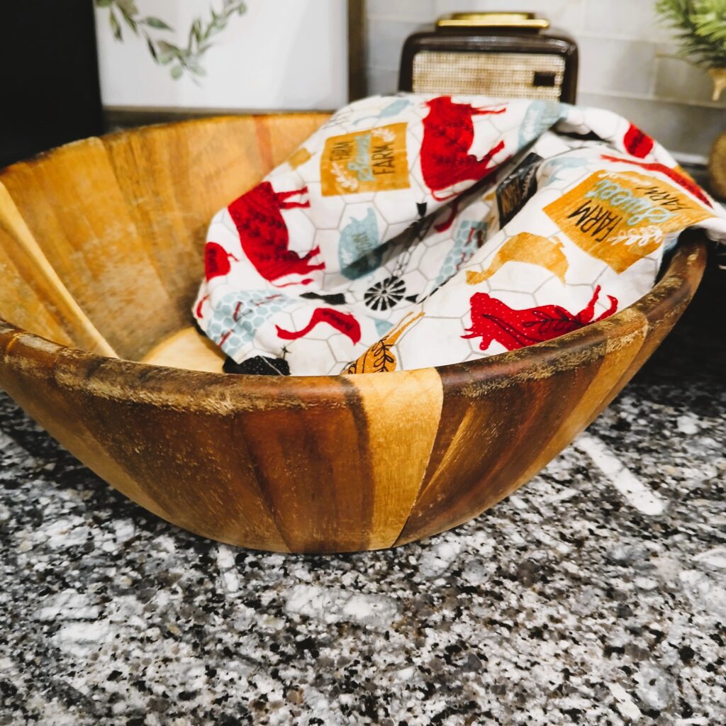 A wooden bowl on a counter with a towel with farm animals draped on the side of the bowl