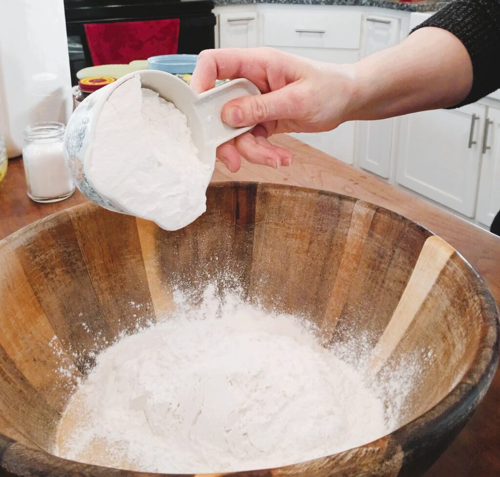 A cup of flour being poured into a wooden bowl