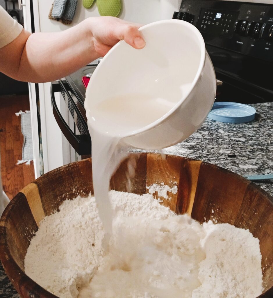 Water and yeast mixture being poured into dough in a wooden bowl